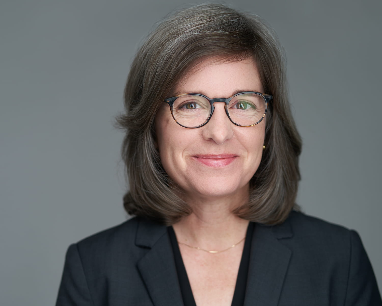 Executive woman in black blazer with glasses - professional corporate headshot against grey backdrop
